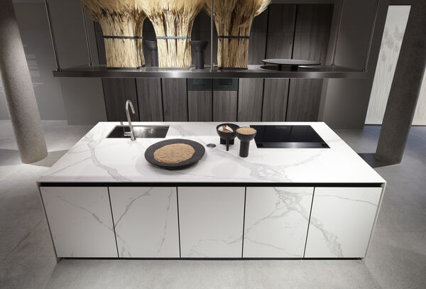 Overhead view of a white marble kitchen island holding a wheat-filled centerpiece, ceramic bowls and a built-in cooktop, with dark wood cabinetry in the background.