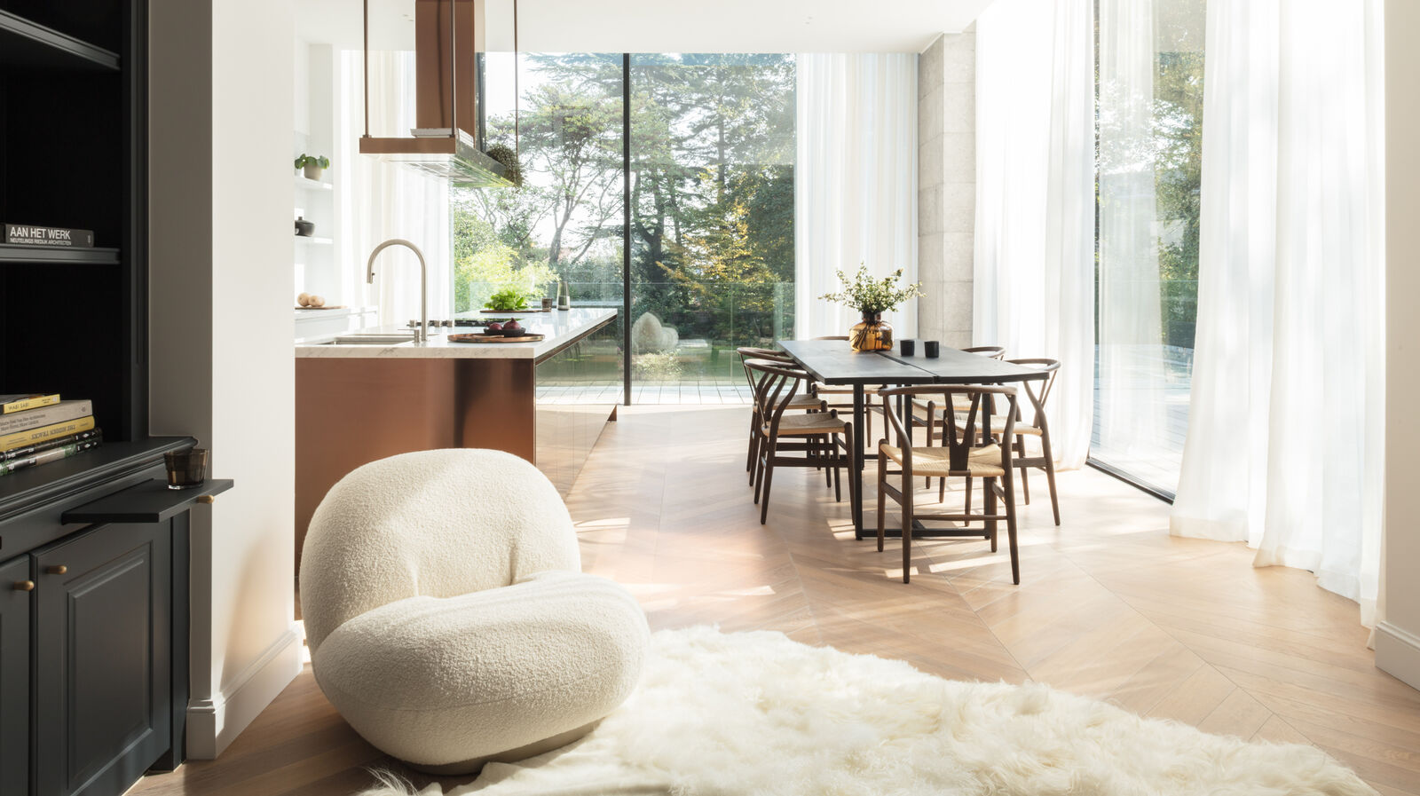 Open-plan kitchen and dining area with a copper-toned island, a cream bouclé lounge chair, herringbone parquet flooring, and full-height glazing onto the garden
