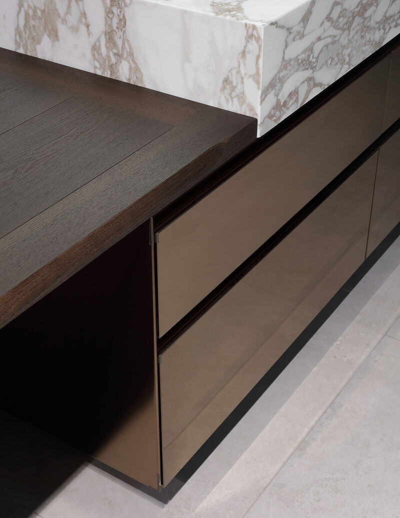 Close up of a veined marble countertop edge above bronze toned glass drawer fronts on a dark wood kitchen island
