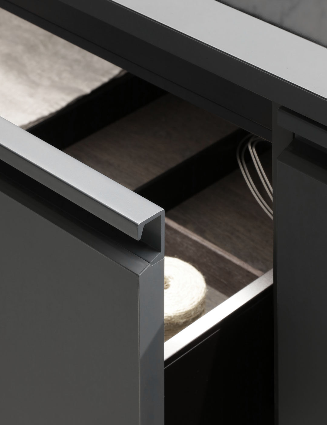 Detail of an open drawer with a slim aluminum handle, showing internal cable management and a rolled linen textile inside a dark wood-lined compartment