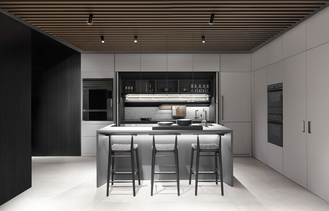 Wide view of a grey showroom kitchen with an illuminated glass-shelf display, marble backsplash, and central island with bar stools beneath a slatted ceiling