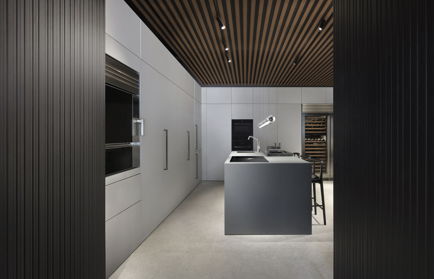 Grey minimalist kitchen with full-height cabinetry, integrated ovens and wine cellar, and a central island, viewed through a dark wood-paneled doorway