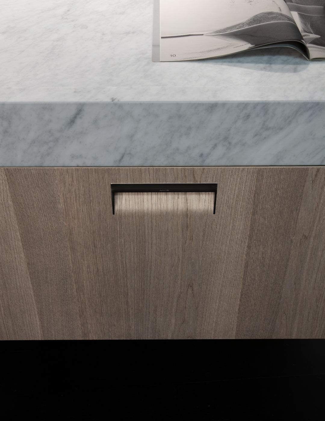 Close-up detail of a marble countertop edge and a wood cabinet drawer with a slim black handle, with an open magazine resting on top