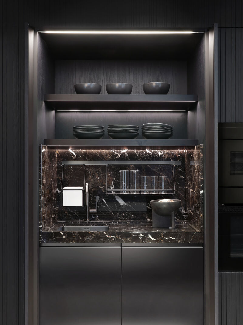 A dark marble backsplash niche with open shelving displaying black ceramic bowls and plates, integrated sink and glass rack in a black kitchen unit