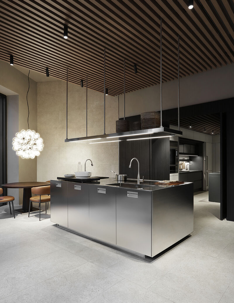 Angled view of a dark kitchen composition with a suspended stainless steel shelving unit, marble worktop, and a round dining table with leather chairs in the background