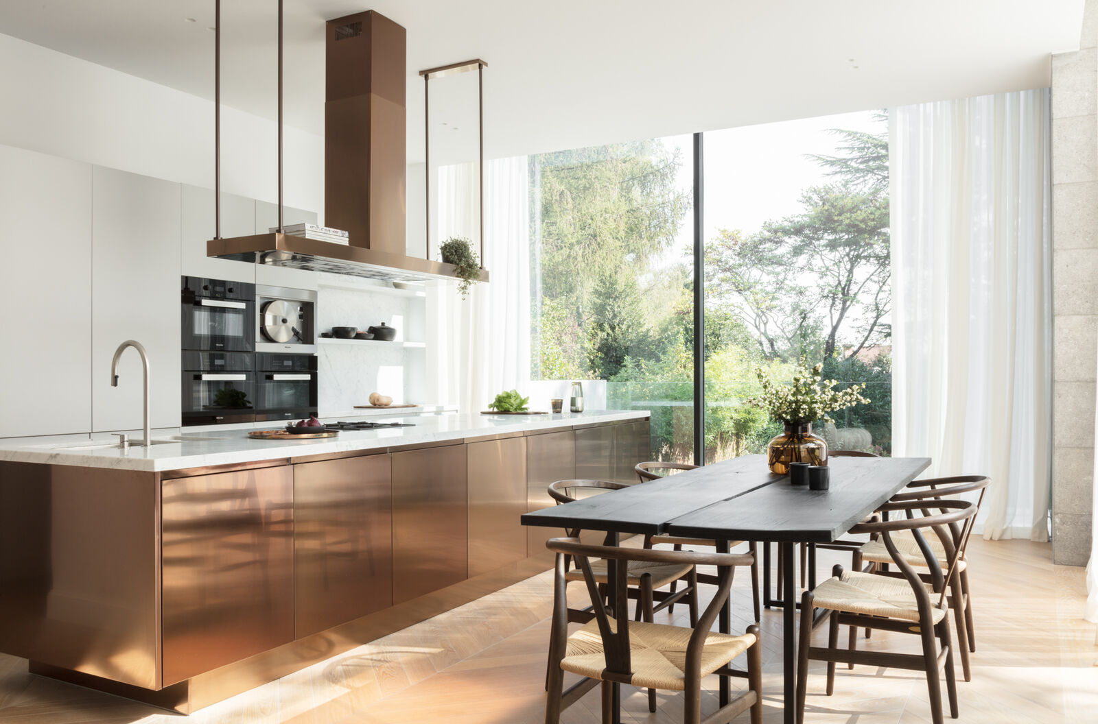 Copper-clad kitchen island with a suspended bronze extractor hood, integrated ovens, open shelving, and a dark wood dining table with wishbone chairs beyond