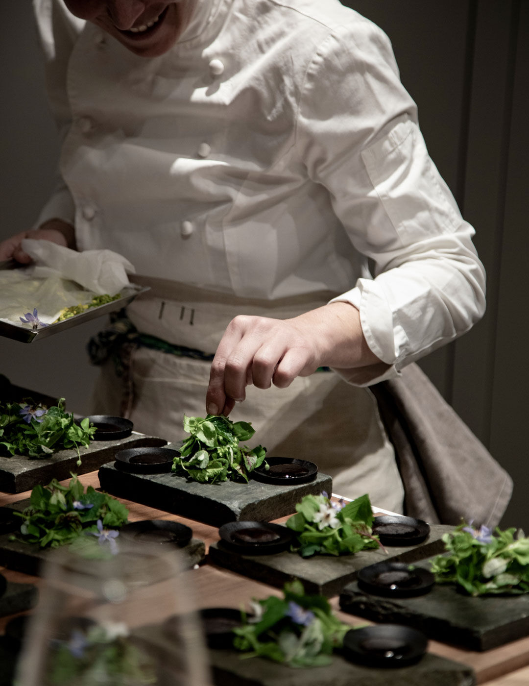 Guests seated around a kitchen island react warmly as one gestures mid-conversation, with stacked plates and glassware filling the counter