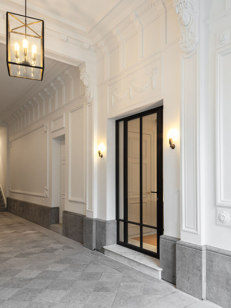 Grand corridor with ornate plaster mouldings, a lantern-style pendant light, a black-framed glass door, and grey stone flooring