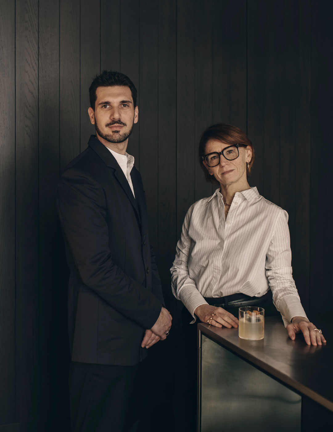 A man and woman pose together at a dark wood-panelled bar counter, with a cocktail garnished with dried citrus resting between them