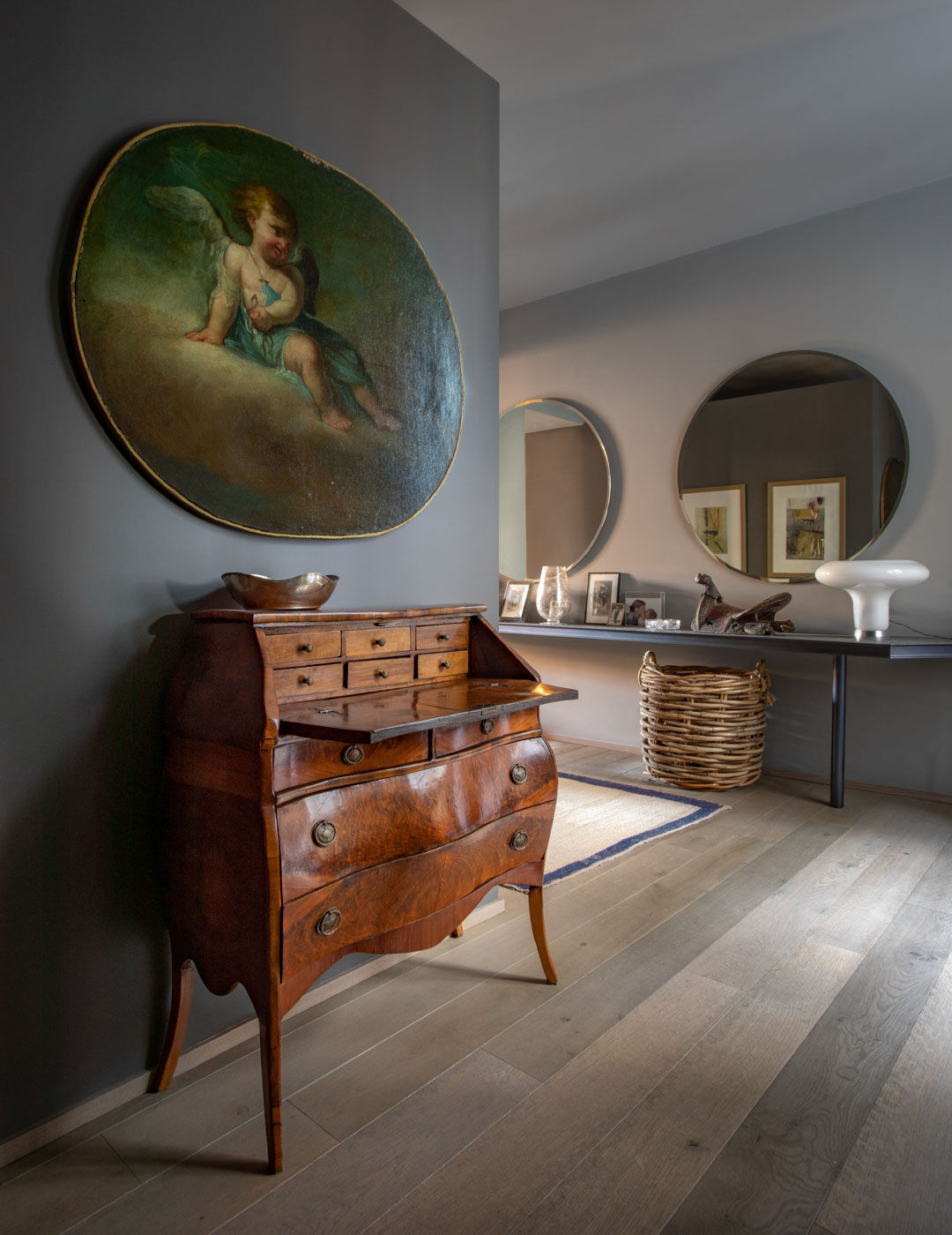 Entrance hall with an antique bureau, oval cherub painting, round mirrors and a wicker basket on wood flooring