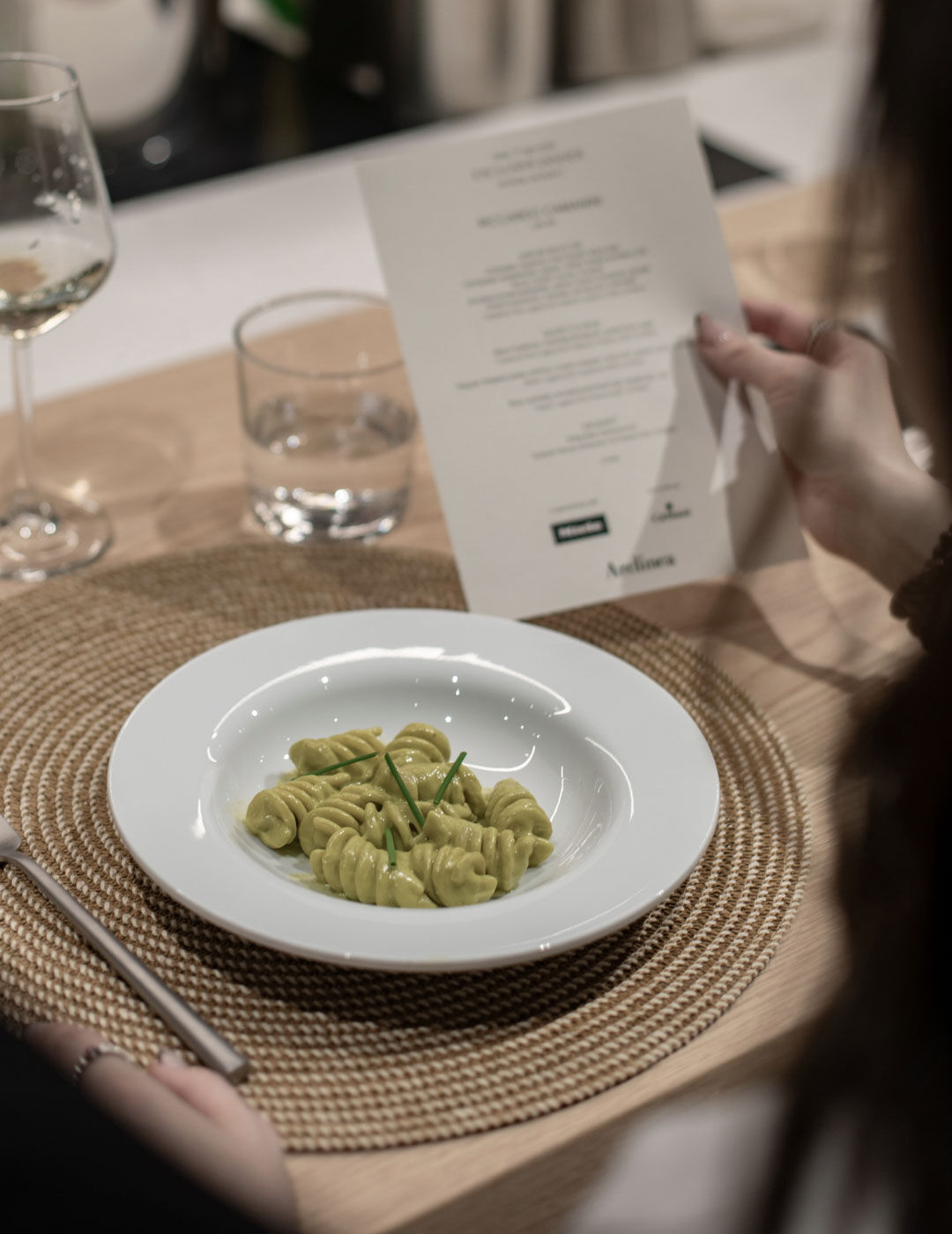 A plate of creamy pistachio-green pasta shells garnished with chives sits on a table beside a dinner menu being read by a guest