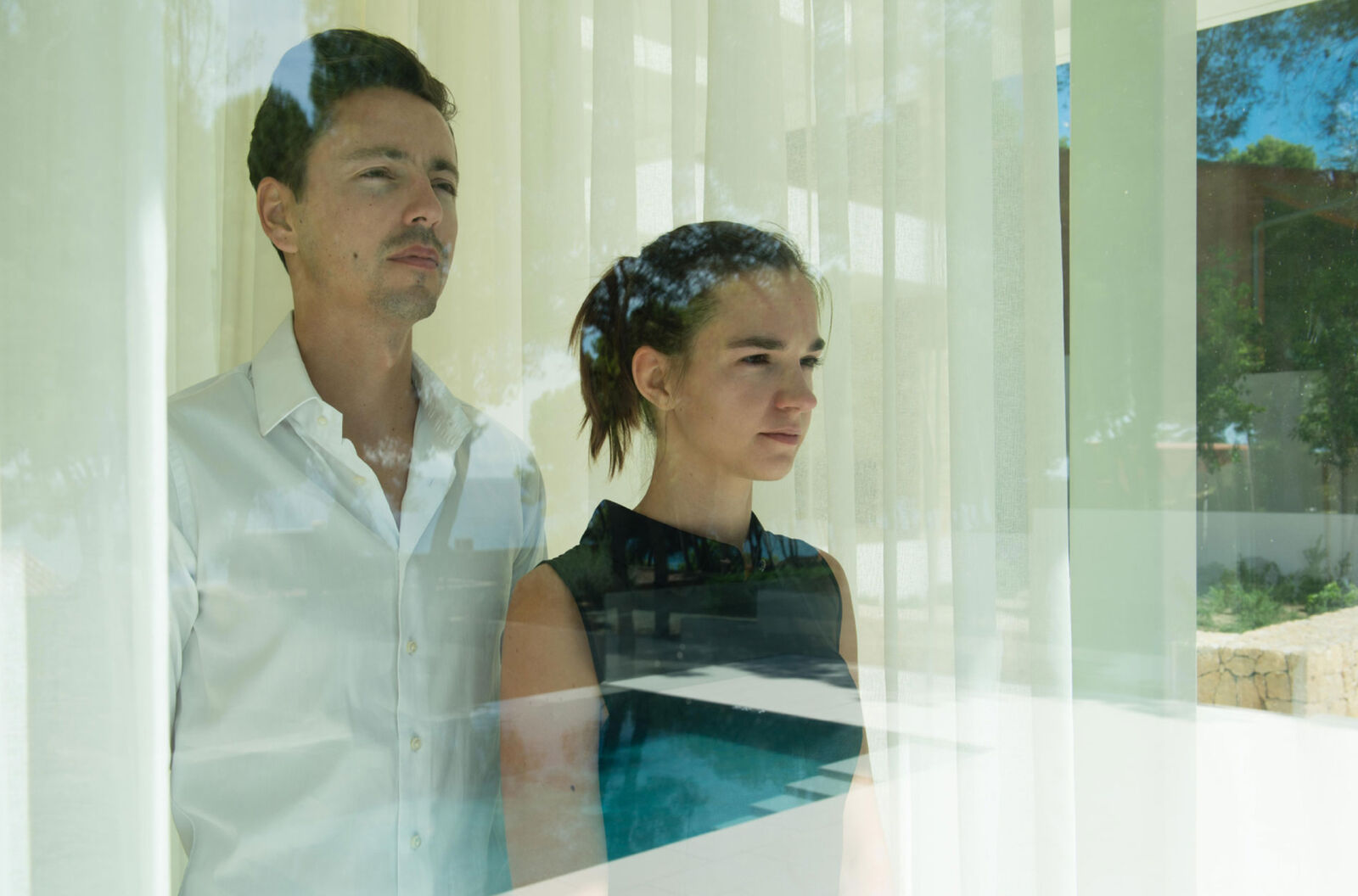 A man and a woman stand close together behind a sheer curtain, backlit by daylight from the garden beyond