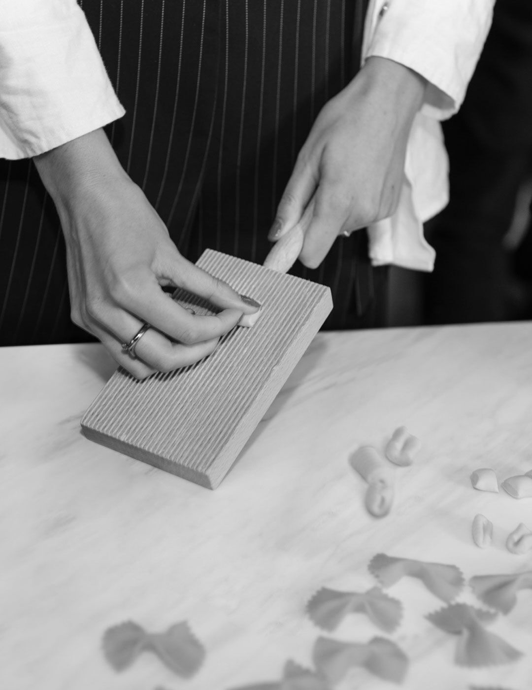 Close-up of hands shaping pasta with a ridged wooden board, with finished pasta shapes scattered on the marble counter