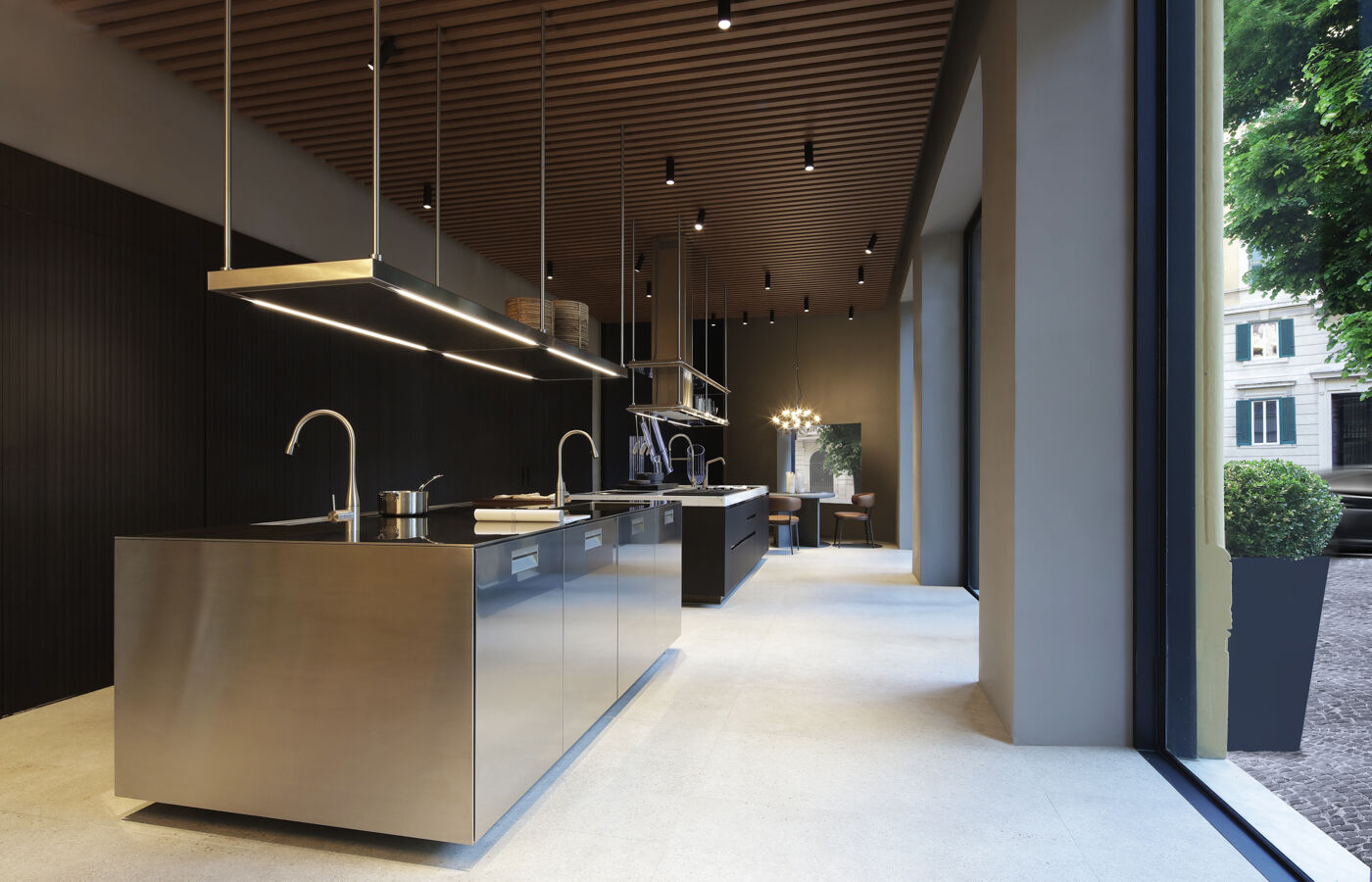Interior of the Arclinea showroom featuring a stainless steel kitchen island beneath a slatted wood ceiling, with a dining area and street view visible beyond