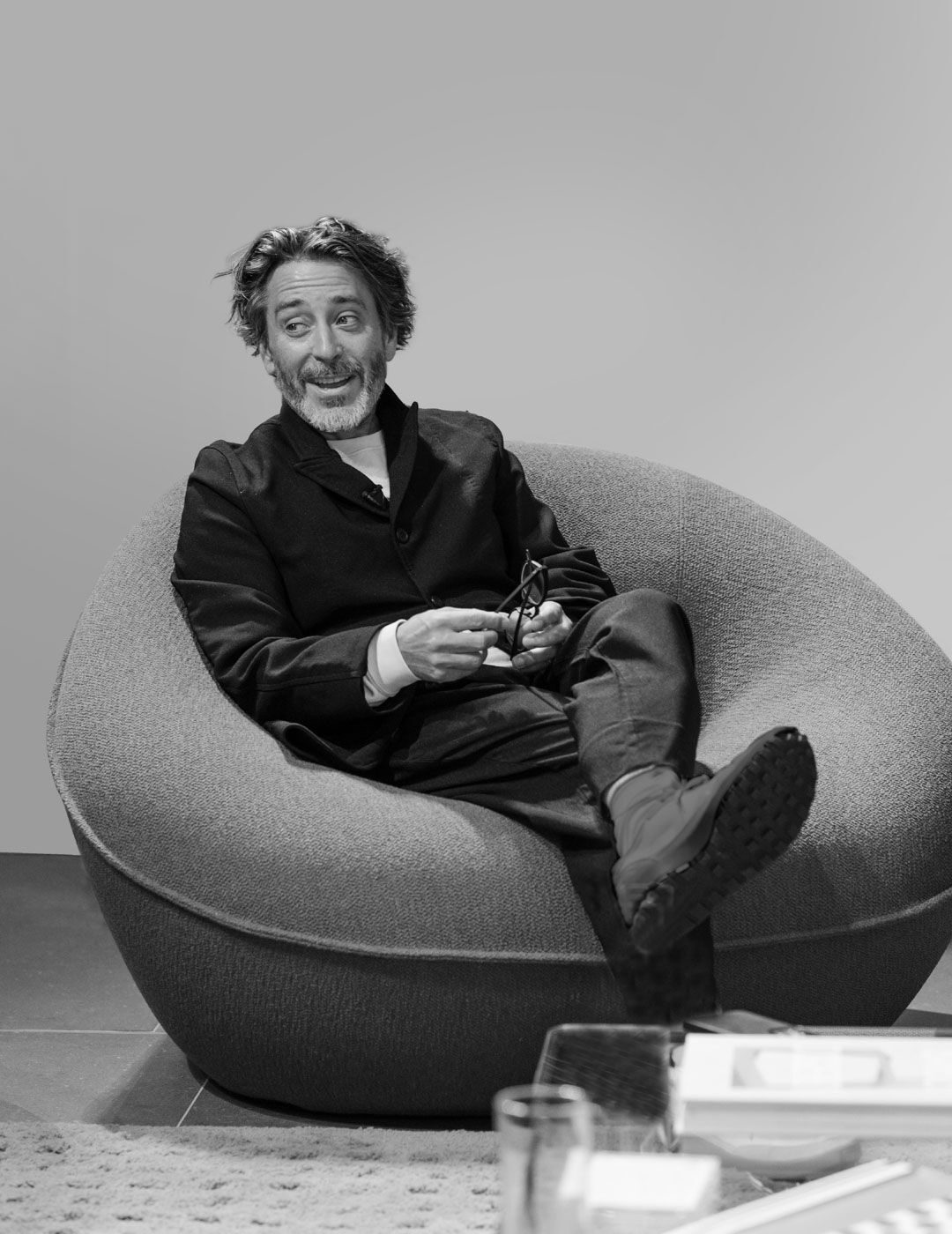 A designer sits relaxed in a rounded upholstered armchair, gesturing with his glasses while speaking during the Brompton Road opening event
