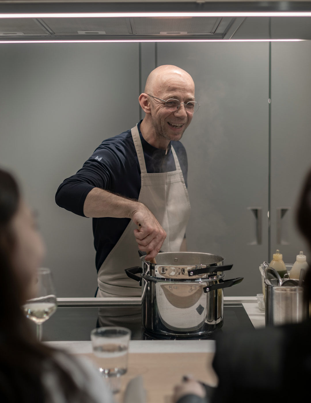 A smiling bald chef in an apron stirs a steaming pot on an induction cooktop as seated guests look on