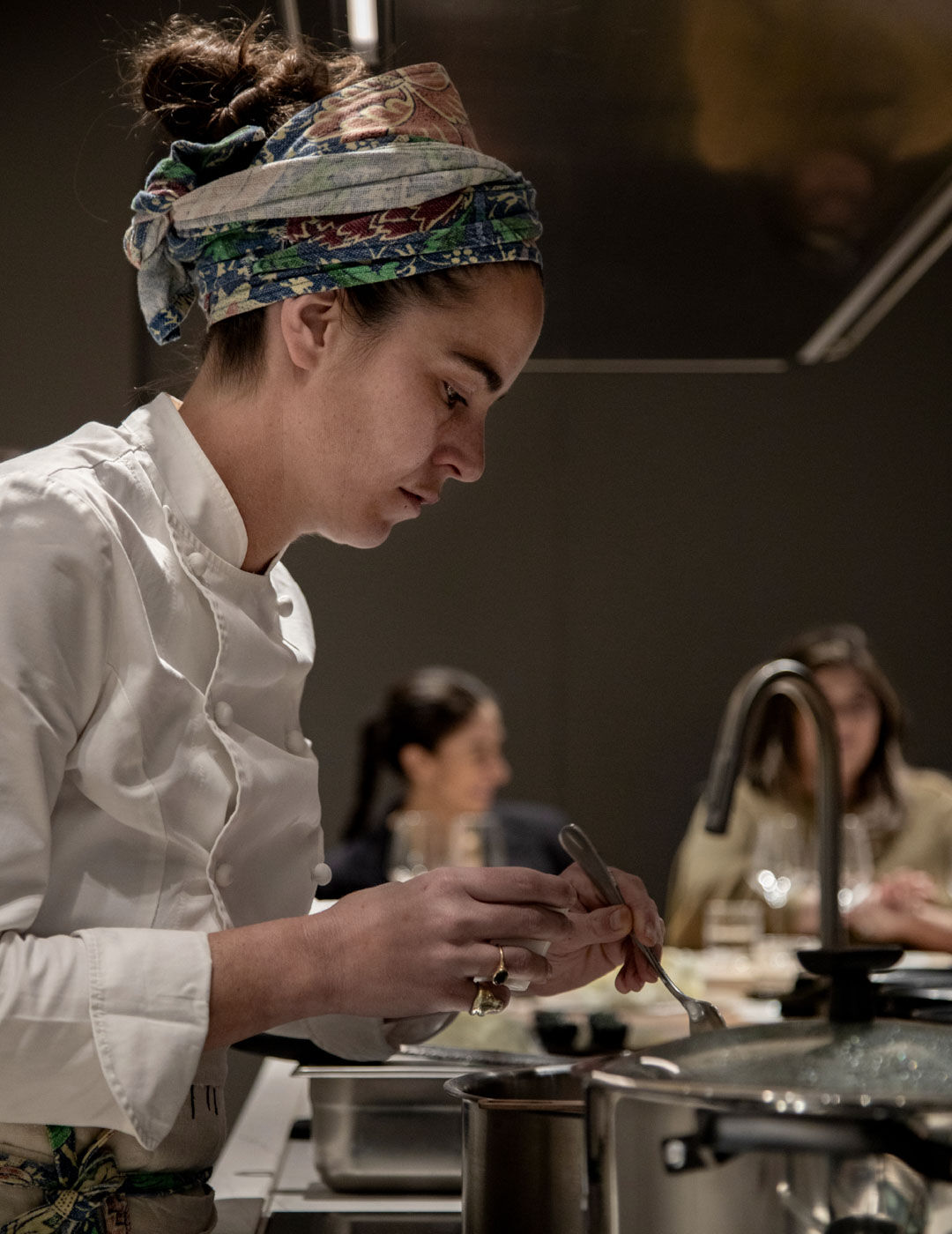 A chef in a floral headscarf carefully seasons a dish with a spoon at the counter as other guests converse in the background