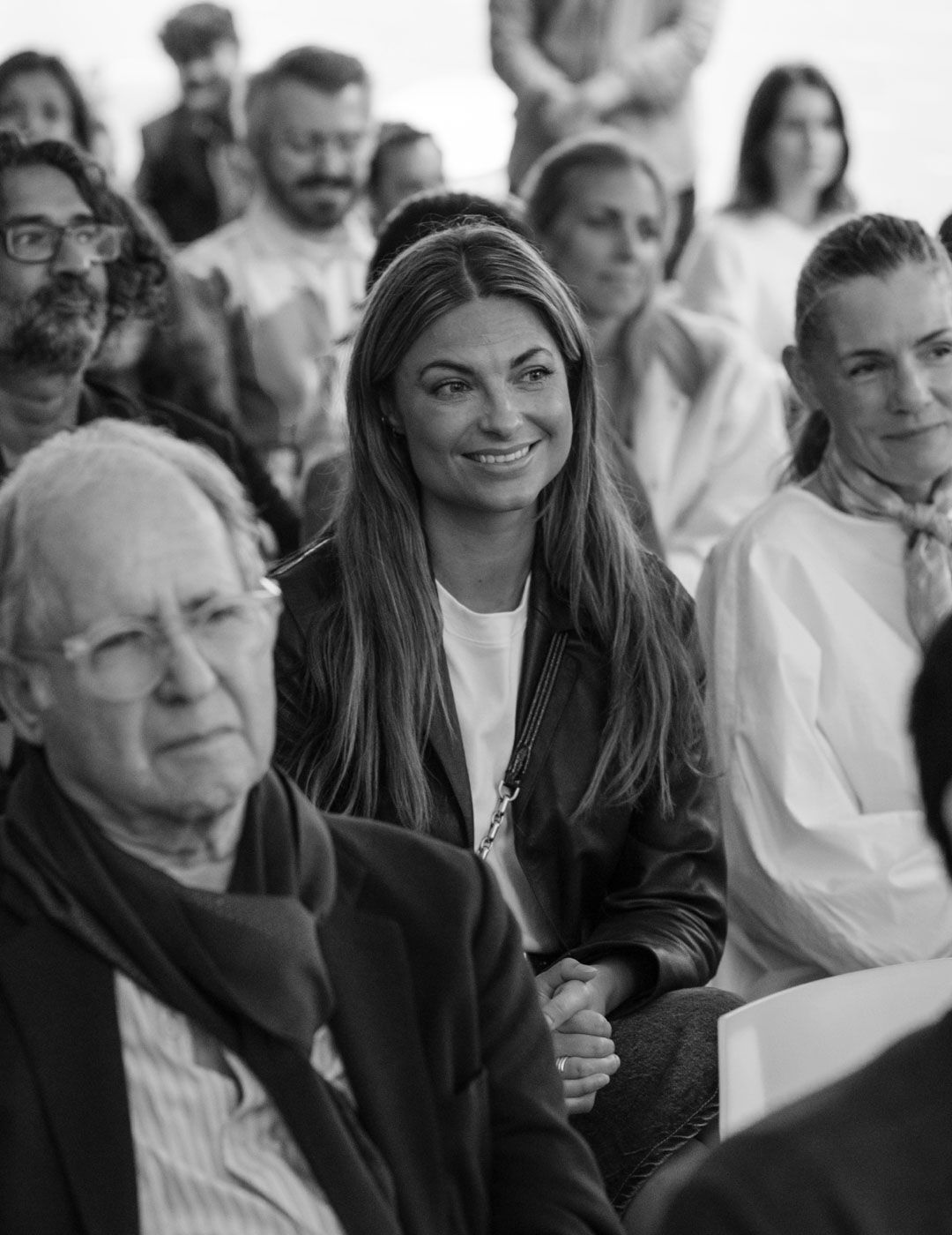 A smiling woman sits among an attentive audience listening to a presentation at the Brompton Road opening event