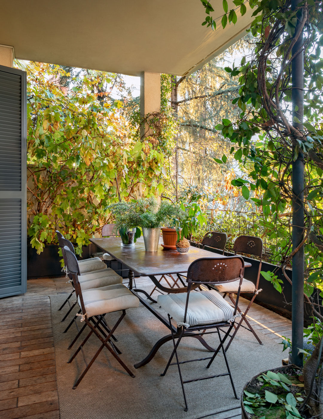 Covered terrace with a rustic table, folding chairs and cushions surrounded by dense greenery
