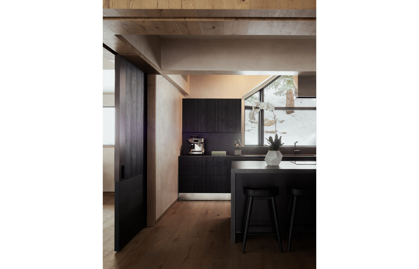 Arclinea kitchen workspArclinea kitchen with black marble island, bar stools, black ash wood finishes, and natural light from a snowy windowace with black ash wood cabinetry, coffee machine, black marble island