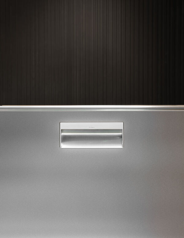 Close-up of a satin stainless steel cabinet front with an integrated recessed handle, set against a dark ribbed wood panel background