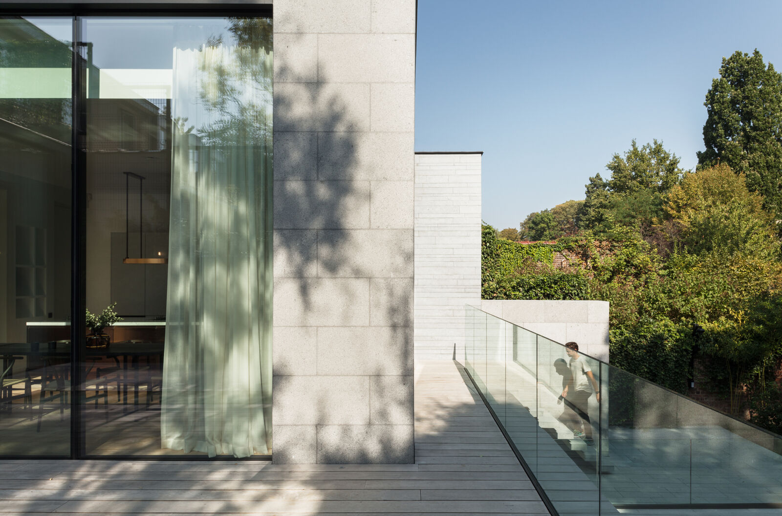 Exterior glass balcony with sheer curtains visible through the window, dappled tree shadows on a limestone facade, and a person walking past a glass balustrade