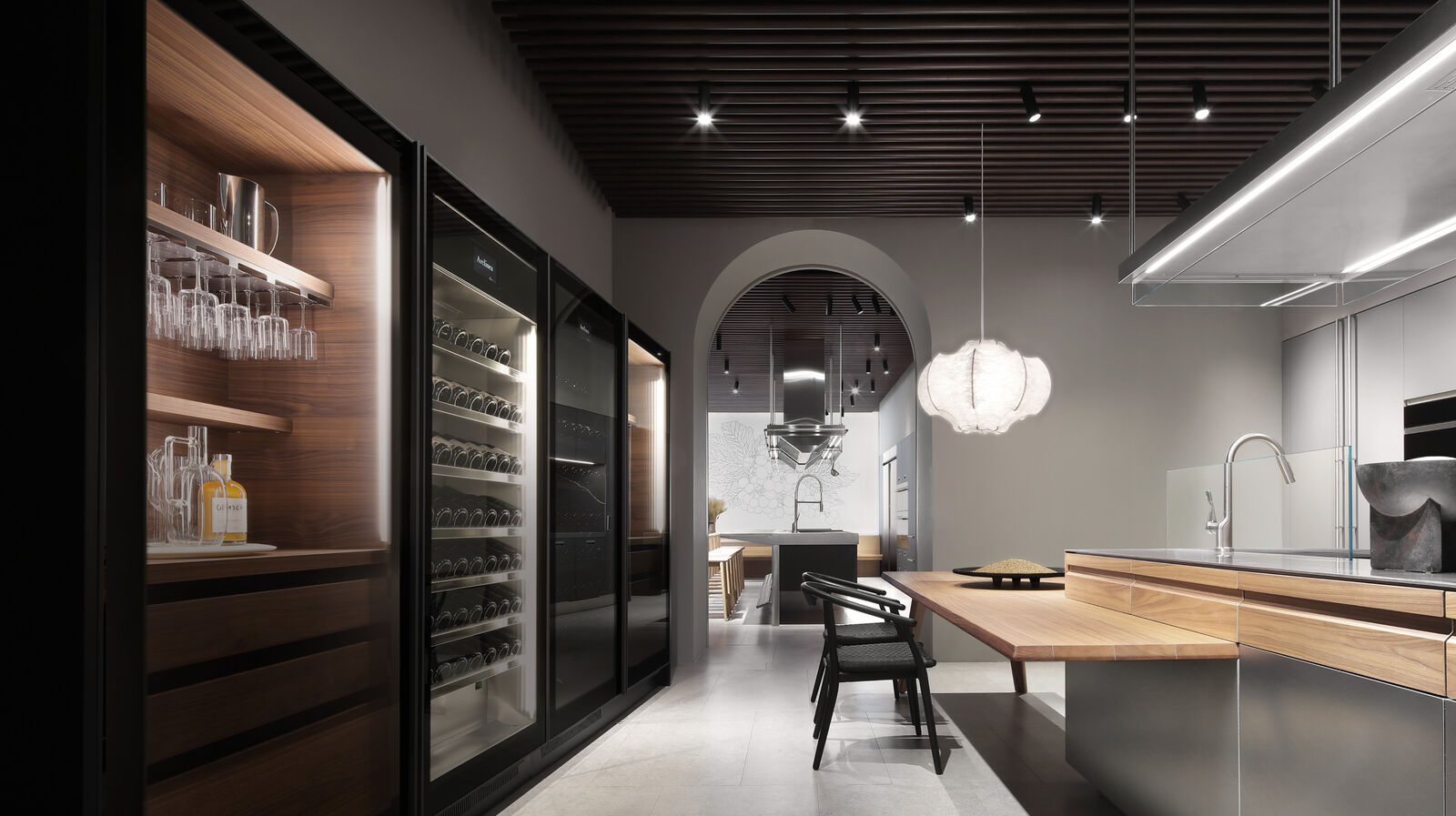 A glass-fronted wine and glassware storage unit with warm wood interior beside an archway leading into a showroom kitchen with a marble island and cluster pendant light