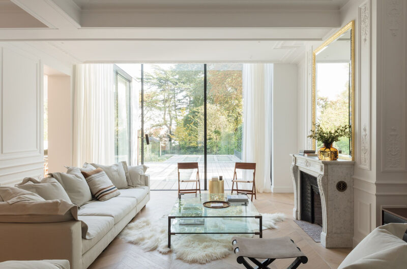Elegant living room with a beige sofa, a glass coffee table over a sheepskin rug, a marble fireplace with gilt-framed mirror, and full-height sliding doors to a terrace