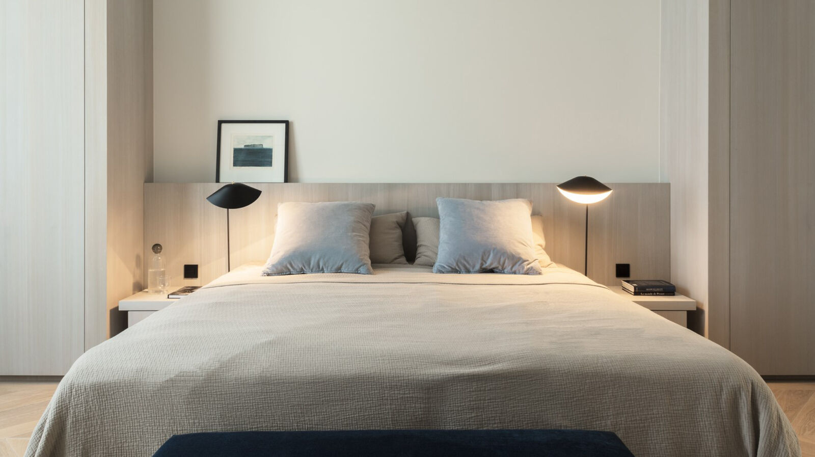 Bedroom with an upholstered headboard, grey linen bedding, matching black reading lamps on floating nightstands, and a small framed artwork
