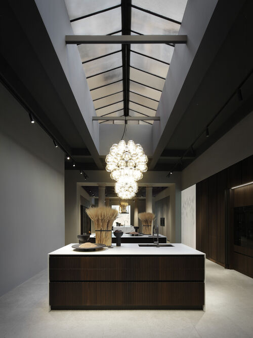 A glass-roofed showroom with a dark wood kitchen island beneath a cluster of spherical glass pendant lights
