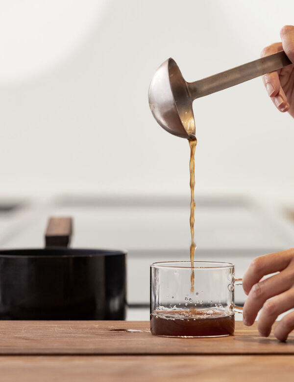 Coffee being poured from a small ladle into a glass cup on a wooden counter, with a dark pot visible in the background