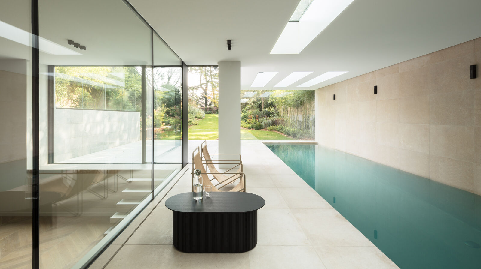 Indoor swimming pool with skylights, floor-to-ceiling glazing onto the garden, and a lounge area with a low dark table and gold-framed chairs