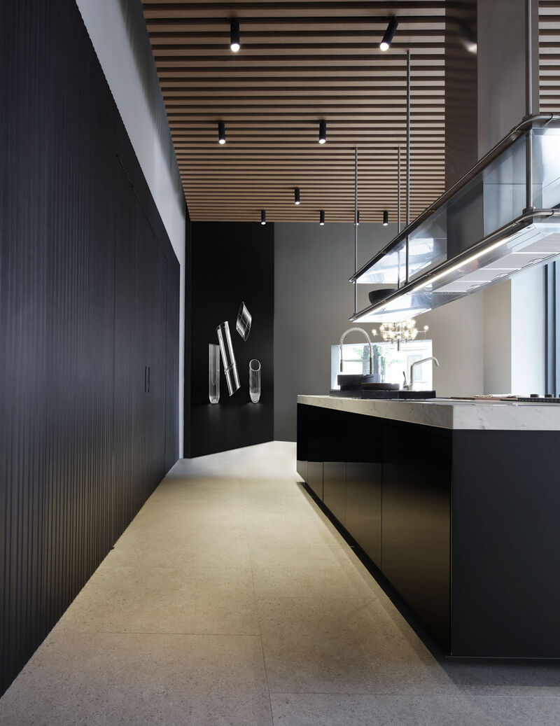 Long view down the showroom corridor past dark ribbed cabinetry and decorative glass objects toward a marble-topped kitchen island with a suspended steel hood