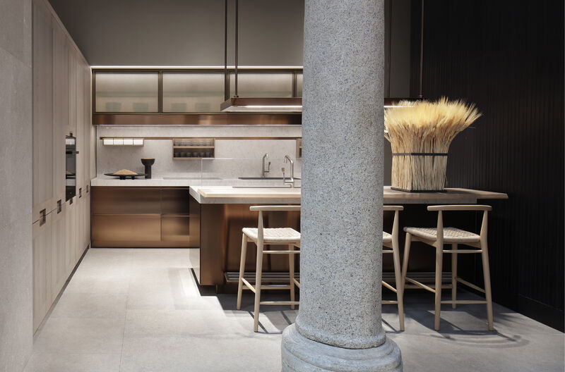 A showroom kitchen with bronze-toned cabinetry, glass-fronted upper units and a stone column standing beside a wooden countertop with wheat-sheaf decoration and two bar stools