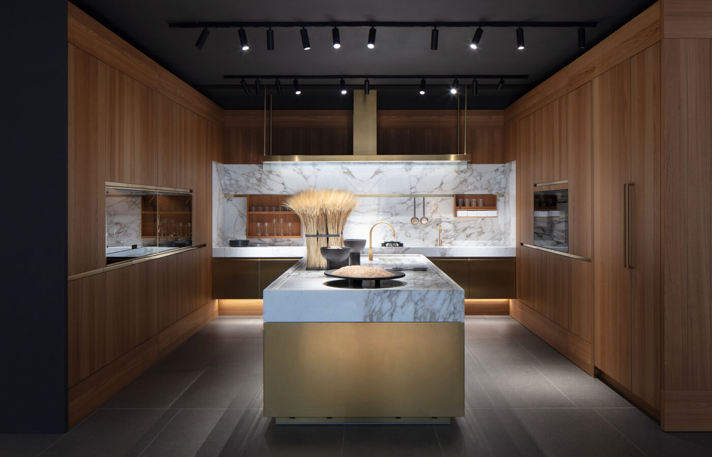 A warm wood-toned Arclinea kitchen with a brass-clad island, marble countertop and brass rangehood, flanked by matching brass cabinet trims