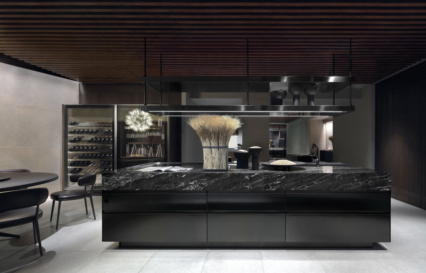 A dark kitchen island with black marble countertop faces a built-in glass wine cabinet, with round table and leather chairs in the foreground beneath a wood-slat ceiling
