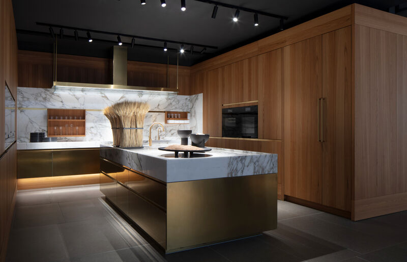 A brass-fronted Arclinea kitchen island with a marble top and wheat-sheaf display, set beneath a matching brass rangehood in a wood-lined showroom