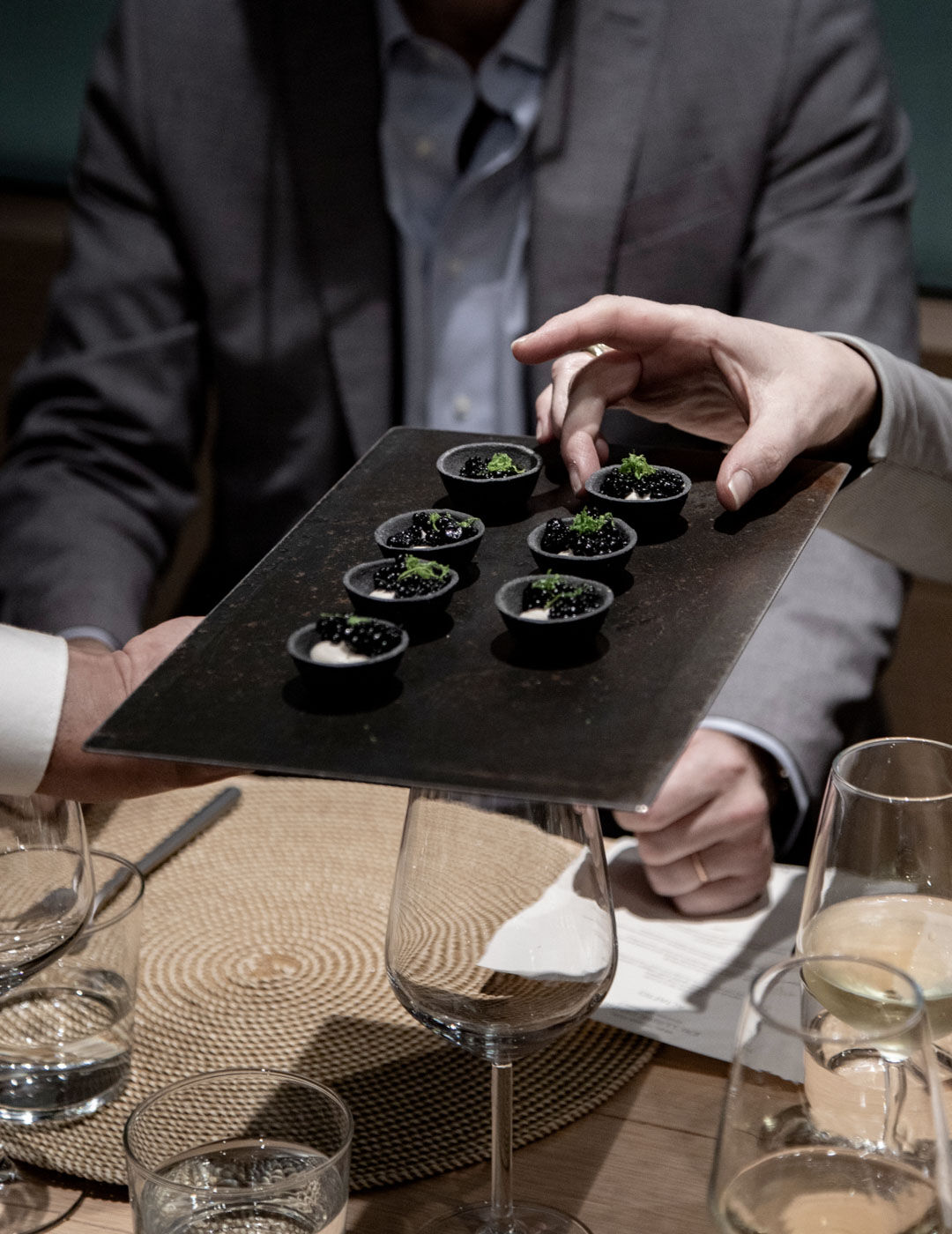 A server presents a slate tray of small tart shells topped with blackberries and mint to guests seated at a table set with wine glasses
